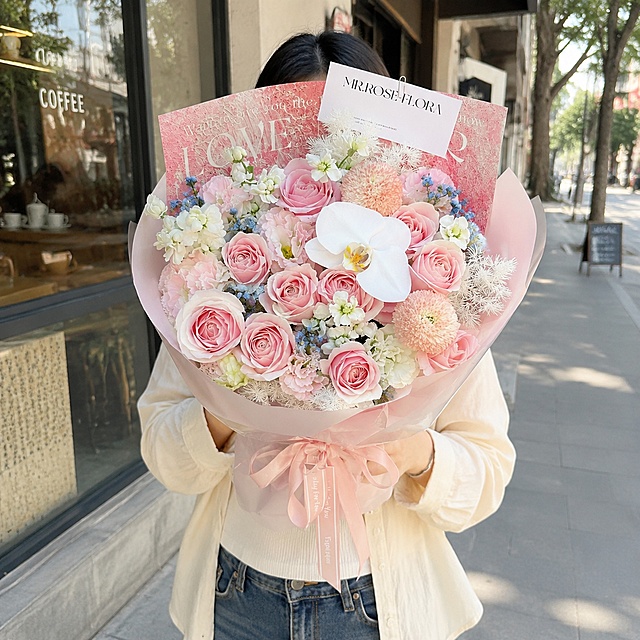 以图1的花束为主体，严格保留花束中所有原有花材（粉玫瑰、白蝴蝶兰、粉色乒乓菊、白色小花、粉色包装纸、LOVE卡片、MR.ROSE FLORA卡片等）的种类、数量、颜色、形态、位置和包装完全不变，仅在花束间隙中少量点缀添加几支图2中的蓝星花（蓝色五瓣小碎花带绿叶），稍微点缀即可，不要加多。去除原图中的人物和汽车背景。画面为白天城市街拍场景，一位中国年轻女性正面抱着这束花，不露脸（头部被花束遮挡或裁切在画面外），自然站立姿态，身穿简约日常服装。背景为白天明亮的城市街道街景，有咖啡店橱窗、人行道、柔和阳光，背景适度虚化突出花束。整体风格清新自然，写实摄影质感，花束为视觉焦点。严格锁定宽高比为1:1。