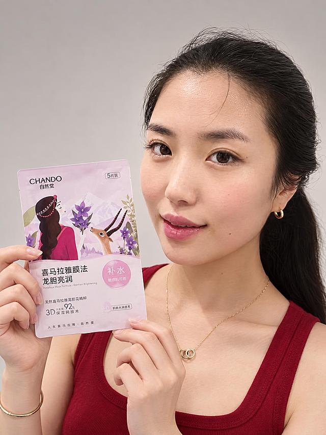专业美妆产品棚拍，年轻东亚女性模特，黑色头发扎成低马尾，面容与参考图中女孩完全一致（严格保留所有面部特征，不做任何修改）。身穿酒红色无袖背心，佩戴金色双环细项链和金色小耳环。模特手持自然堂喜马拉雅膜法面膜袋展示，微笑表情。面膜袋为龙胆亮润款，淡粉色包装，插画女孩（玫红衣服、珠饰发带背影）、藏羚羊（gazelle造型）、紫色龙胆花，文字清晰：喜马拉雅膜法 龙胆亮润 补水 敏感肌可用 92% 3D保湿网技术，包装细节与参考图完全一致。纯色中性浅灰米色渐变影棚背景，干净简洁。专业影室柔光，柔和均匀，立体感强。半身景别，正面持产品展示。超高清2K画质，真实摄影质感，皮肤细腻自然保留真实毛孔纹理。双手正常10指。简约高级美妆广告风格。