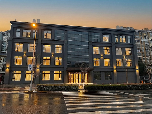 将这张建筑图片的整体风格改成参考图的风格，参考图是偏暖调的湿地面城市街景，现代简约深灰色建筑外立面，傍晚氛围，同时移除图片里所有停放的汽车，移除所有店铺招牌和酒店名称文字，保留建筑整体结构和构图，保持原街道和绿化的布局