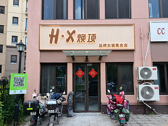 将图中红框位置的门头替换为浅色长城板（米白色凹凸格栅）底板的女装店门头，四周加精致玫瑰金金属边框，门头文字内容为"H·X 焕顶 品牌女装集合店"，文字采用玫瑰金立体字背发光黄色暖光效果，字体优雅高级，整体风格轻奢简约，符合高端女装店气质，门头安装在粉色建筑立面上，白天自然光效果，真实感强，高清细节