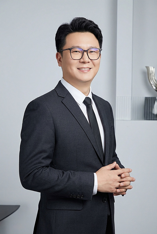 保持原人物脸部五官一致，生成职业写真照。人物身穿剪裁合体的深色职业正装，面带自信微笑，正对镜头，双手自然交叠在身前，姿态自信大方。上半身特写，略带一点角度的半身构图，符合商业人像三分法构图原则。摄影棚灯光，背景为浅灰色带简单装饰，整体画面干净干练。保留人物原有的眼镜。