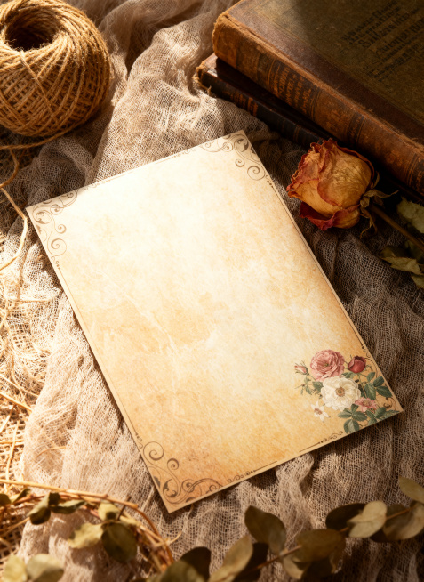 超写实静物摄影，俯拍 45° 视角，必须严格保留参考图里A4信纸的样式（复古泛黄底色、卷草花纹边框、右下角花卉图案），将这张信纸整体平铺在亚麻纱布上；画面右侧点缀干枯玫瑰花，复古书本，左上角放置一团天预览效果