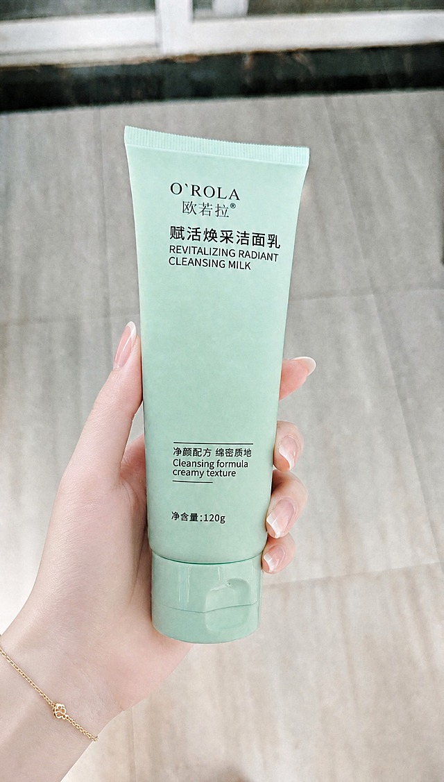 参考第二张图中女性手部的样式，将第一张图中拿着洁面乳的手替换为同款精致白皙的女性手部：手指纤细修长，皮肤白皙细腻，指甲为自然裸色法式美甲，手腕佩戴同款细款金色小花手链。保持握持洁面乳的姿势自然，产品大小、位置、所有文字内容和背景环境完全不变，整体光线、阴影和色调与第一张图一致，真实自然的高清摄影质感。