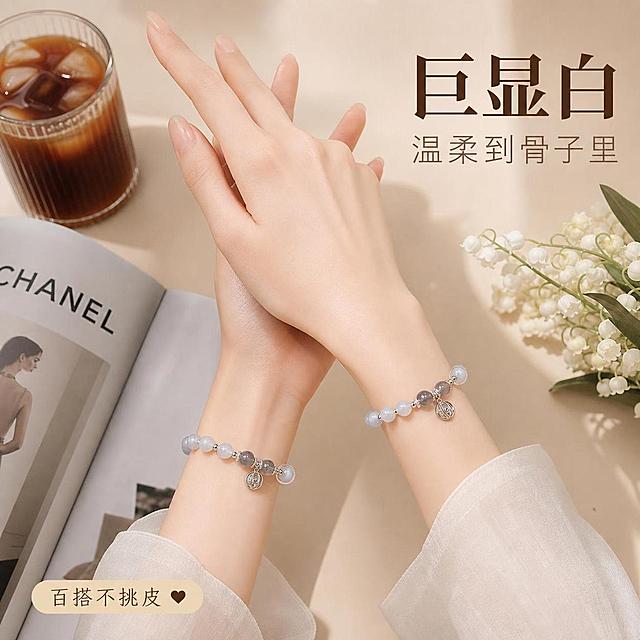 [主图-4] 手串佩戴效果展示 显白显气质 多场景穿搭
[氛围风格] 温柔高级日常感 轻奢生活方式
[画质] 高清生活方式摄影 光线自然柔和 画面干净有格调
[元素] 第一视角双手自然交叠于身前 两只手腕都佩戴着手串 展示不同角度的佩戴效果 手部皮肤白皙 手指修长 背景是浅米色高级书桌 上面放着一杯冰美式咖啡 一本打开的时尚杂志 一小束白色铃兰花 整体营造精致女生日常氛围感 保持手串外观（即物理结构 材质质感 颜色 珠子排列 吊坠造型）与输入产品图一致 杜绝任何变形和色差 精细化处理 光影随场景自然融入
[光影] 窗边自然光主光 柔和漫射 从左侧照入 手部有自然明暗过渡 珠子有温润高光 整体暖调柔和
[配色] 主色温柔米杏色 辅色银色珍珠白 强调色淡粉肤色
[视角构图] 第一视角俯视 双手位于画面中心 桌面场景丰富但不抢主体 中等景深
[文字版式] 右上大字"巨显白" 粗体深棕色 副标题"温柔到骨子里" 小号浅棕色 左下"百搭不挑皮"米色底深棕字小标签 配小心心图标
[输出要求] 1:1比例 文字清晰可辨 超高清生活摄影质感
[负向提示词] 变形 文字错乱 产品模糊 背景杂乱 水印 电商平台Logo 多指 手指畸形 手部扭曲