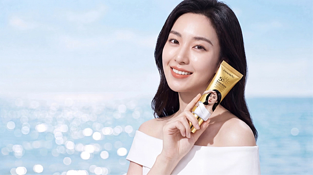The camera slightly pushes in. The woman smiles radiantly and confidently into the lens while elegantly holding the gold sunscreen tube near her face. The product in her hand remains perfectly stable and clear. The bright summer sunlight beautifully illuminates her perfectly protected flawless skin. In the background, the light-blue ocean ripples and sparkles brilliantly. High-end cosmetic commercial ending shot.
