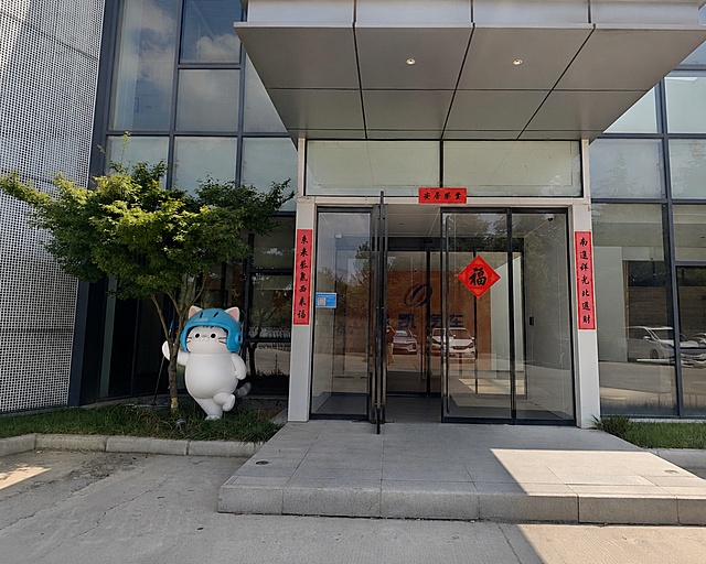 保留原图片公司入口外立面建筑、春联、福字和logo不变，将带蓝色头盔的白色冠军猫IP添加在门口树木旁，冠军猫单腿支撑，一只胳膊贴在树干上，整体呈依靠树干姿态，正对镜头，画面真实自然融合
