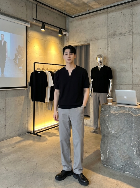 侘寂工业风男装店内，裸露水泥毛坯墙搭配水泥地面，粗粝极简氛围。左侧带暖光灯黑色挂衣杆挂着黑白简约成衣，墙面投影幕布播放时装秀。银色金属无头模特穿同款黑色短袖V领上衣和灰色宽松直筒裤。右侧石质台面放笔记预览效果