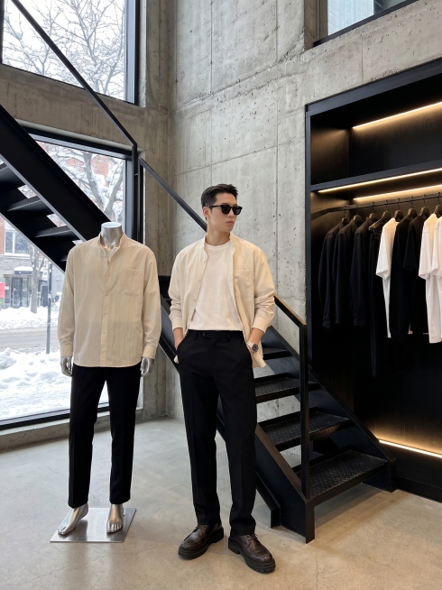极简工业风男装店，氛围高级清冷，黑色钢结构楼梯贯穿画面，裸露水泥墙面线条硬朗。右侧内嵌暖光灯黑色挂衣区整齐陈列黑白简约成衣，落地窗外冬日街景树木建筑带积雪，冷调自然光与店内暖光对比。旁边立着银色金属无预览效果