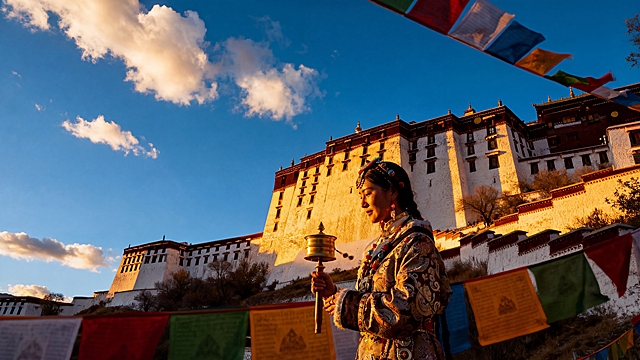 在画面前景增加一位穿着藏族传统服饰的女性，手持转经筒，保持原有布达拉宫、飘扬的经幡、金色夕阳、蓝天白云的场景，电影级画质，旅行大片风格，光影质感高级