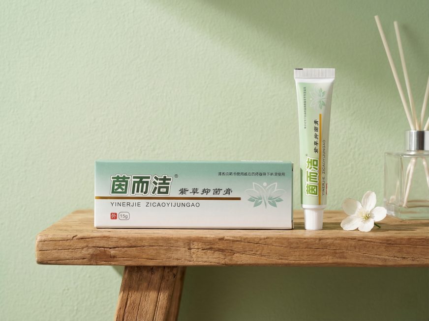 保持茵而洁紫草抑菌膏包装盒和软管的所有文字、Logo、外形完全不变，产品放置在原木浴室洗手台搁架上，旁边搭配一朵素雅的白色小花和透明玻璃香薰瓶，背景是干净的浅绿调硅藻泥墙面，柔和的自然光从侧面照入，营预览效果