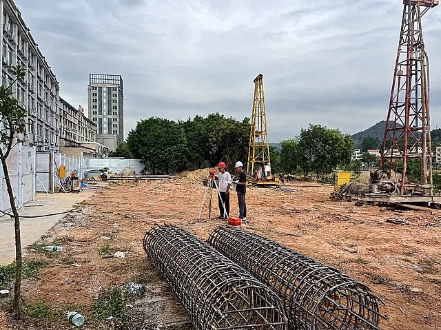 将图中的两个钢筋笼移动到一起，位置往前（靠近画面前景方向）移动，只露出一半（后半部分被画面底部边缘裁切），两个钢筋笼并排靠在一起放在画面下方偏中间的泥土地面上。保持钢筋笼的真实质感、光影、阴影与原图阴天环境一致，与地面接触自然，其余场景元素和人物保持不变，整体写实摄影风格不变。