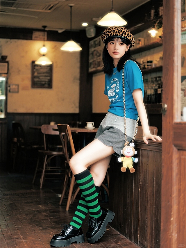 在复古咖啡馆内，潮流女孩戴豹纹贝雷帽，穿湖蓝印花短T、灰色短裤，配黑绿条纹长袜与厚底鞋，背链条装饰包包，挂萌趣公仔，慵懒倚靠，灯光打造时尚个性感