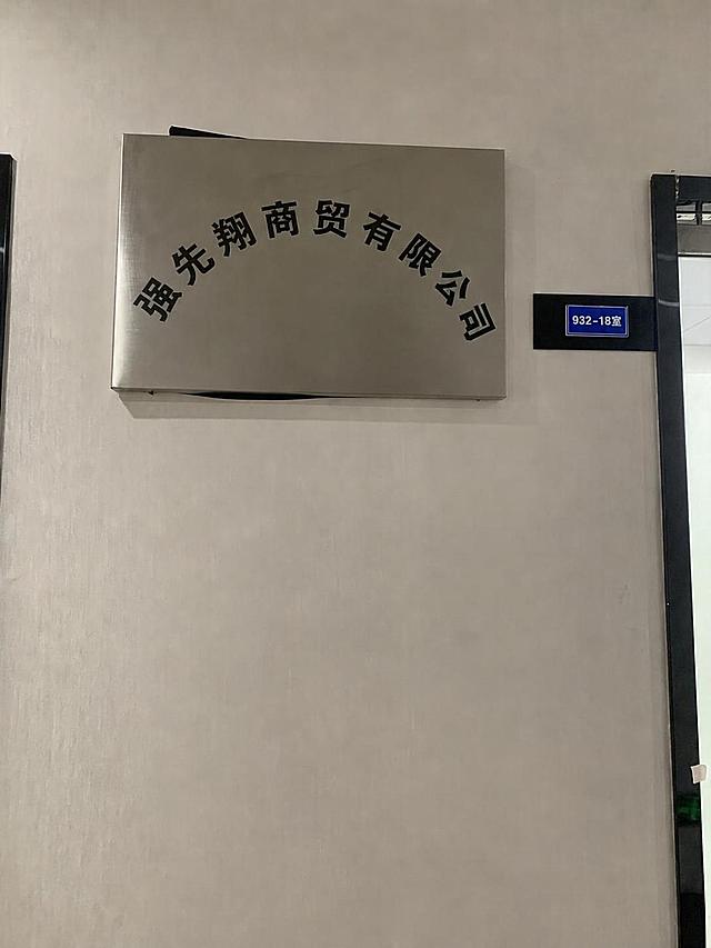 将图片银色公司门牌上的原文字“圣方庆商贸有限公司”改为“强先翔商贸有限公司”，保持字体风格、颜色和弧形排版不变，保持门牌整体样式、背景墙面和旁边门牌不变，只修改公司名称文字