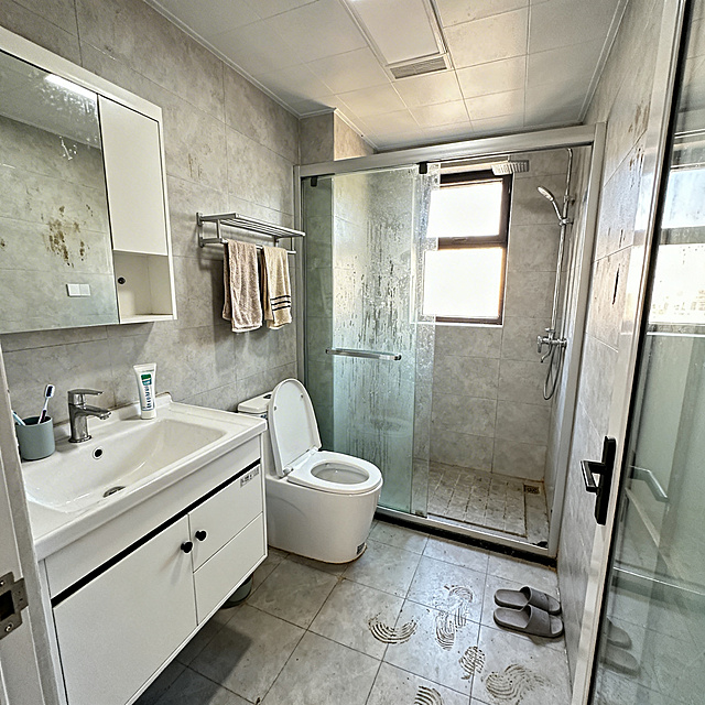 公寓洗手间实拍，干湿分离设计，带有玻璃淋浴房，白色洗手台搭配镜柜，壁挂式马桶，地面和墙面都铺贴浅灰色哑光瓷砖，有一扇小窗户，自然光从窗户照进来，洗手台台面上放着牙刷杯和洗面奶，毛巾架挂着两条毛巾，整体干净清爽，带有一点日常使用痕迹，真实的家庭洗手间场景，无人物，手机实拍效果