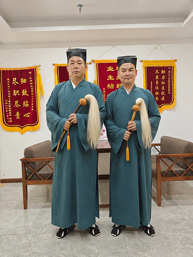将图中两位男士更换为传统道士造型：身穿青色道袍（道士传统服饰），头上佩戴道士巾帽，人物手中拿着拂尘，保持人物的面部、表情、体型、站姿完全不变，背景中的锦旗、桌椅、墙面、地面、天花板等所有环境元素完全保留不变，整体光线、色调、透视与原图一致，真实自然，无PS痕迹