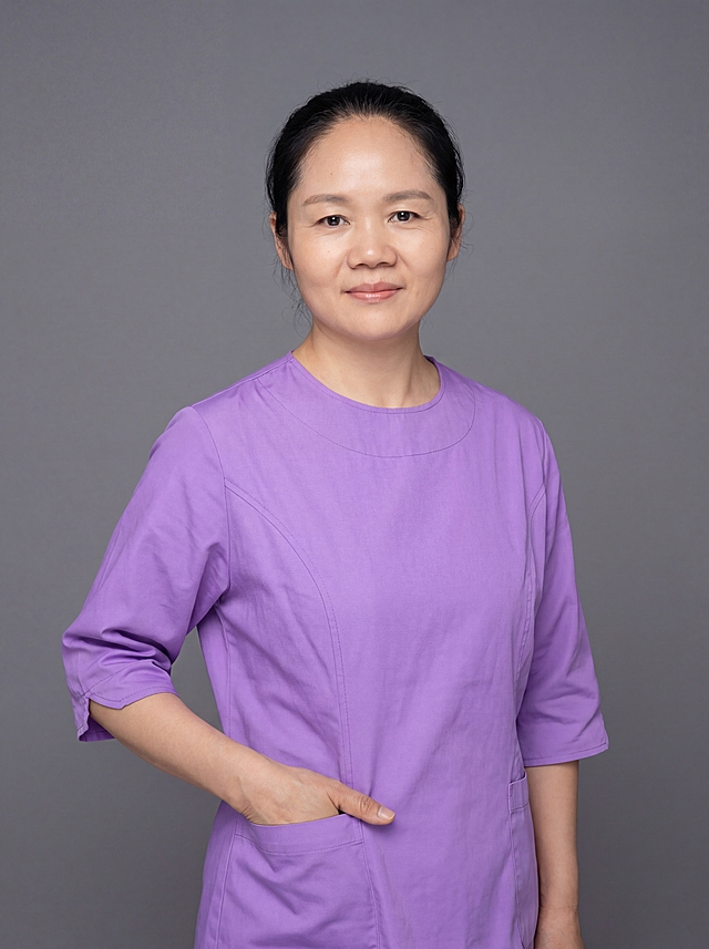 参考图中保留人物脸型、发型、年龄等特征生成医护职业形象照,标准焦段50mm,f/4光圈(f/2.8-f/4区间),平视视角(镜头与人物视线齐平);人物占画面70%,人物居中(居中构图),位于画面中心,为视觉聚焦点,入境范围头部及以下至臀部下方10cm(半身像);左右留少量背景空间;头部与画面顶部保留5%空间;头部发型完整,头肩比例协调,人物结构准确;发质微微柔顺，微微蓬松。身穿紫色医护工作服，高支棉材质，版型整洁得体；着装正式干练，专业温和，身形匀称，姿态端正，身体挺拔。呈自然站姿，左手插兜，肢体放松且自信；去除口罩。简约微微中度灰色纯色背景(呈现均匀柔和质感),背景光线均匀柔和,强化色彩对比的高级感，极简干净;画质超高分辨率、细节清晰,现代精致感风格;肤质精细化处理;眼神直视镜头,目光坚定且澄澈,表情自然，嘴角上扬;清透自然裸妆(轻薄底妆+淡唇妆)妆感轻薄轻透裸肌感,通透感肤质,肤质自然细腻,肤色健康均匀,雾面柔光裸肌感,保留真实皮肤细节｡正面柔光补光,4800K中性光,面部光比1:1.5,色调明亮｡中近景写实摄影,对焦清晰,高清保留皮肤细节｡分辨率4K,去除人物之外的所有元素。严格按照图片内容生成，画面比例3:4