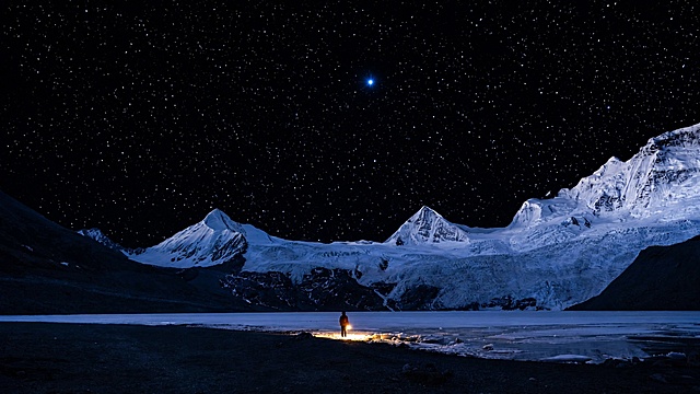夜空繁星点点，雪山静谧壮美，一人立于湖畔篝火旁，画面宁静深远，适合文旅旅游宣传使用