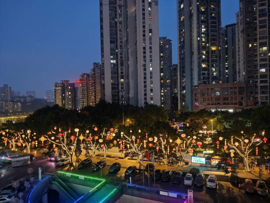 在城市夜景中，对红色框选区域的树木进行文旅亮化设计：树上悬挂迎春灯饰挂件和氛围灯，树干缠绕暖色系LED灯带，灯光效果真实自然，符合实际3D效果。保持原图视角和建筑背景，调整图片角度使其更正，呈现多角度预览效果