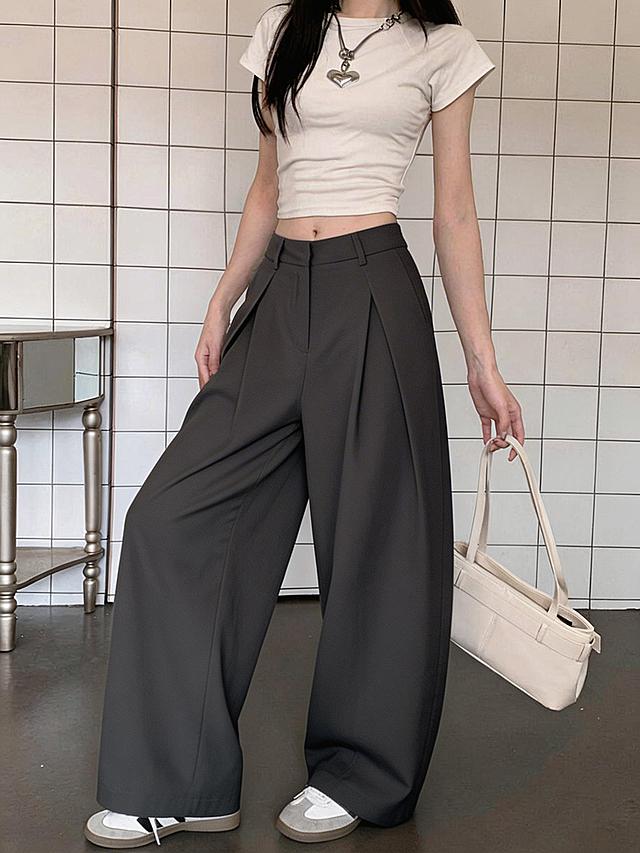 将图二的深灰色阔腿西装裤替换到图一模特的下半身，保持图一模特的姿势、白色短袖上衣、银色心形项链、手提的白色包包、背景方格瓷砖墙、银色边桌、灰白色运动鞋完全不变，裤子版型完全还原图二的褶皱设计，光影与整体画面融合自然，无违和感