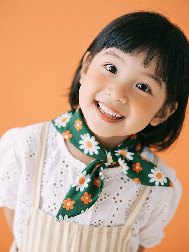 明亮的纯橙色背景，一个穿着白色镂空花纹衬衫和米色条纹背带裙的小女孩，脖子上系着一条绿色底印有白色雏菊和橙色小花图案的丝巾，她微微歪着头，睁大眼睛，嘴角上扬，露出开心的笑容，眼神中带着一丝俏皮和好奇，整体画面色彩明快，光线柔和，人物表情生动自然，具有清晰的肖像摄影风格。