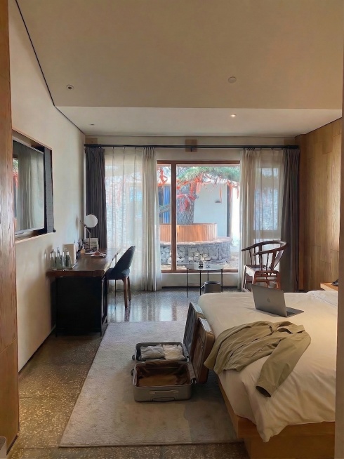 在床靠近行李箱的位置放置一件浅卡其色薄外套，自然搭放；在外套旁放置一台打开的银灰色笔记本电脑，自然摆放，不遮挡行李箱。保持原图整体新中式禅意民宿结构、光影、地毯上的打开行李箱和其他所有物品不变。光影匹预览效果