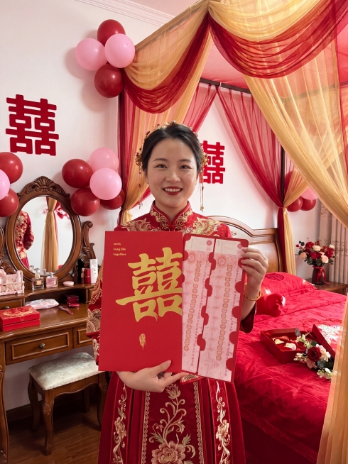 画面中心为身着红色中式传统礼服的人物，左手托住红包底部，右手扶在红包右侧边缘，将红包举在胸前，面部完整露出，笑容自然。 红包为左右两页展开式设计，占画面核心视觉区域近 50% 面积，保持红包原有样式不预览效果