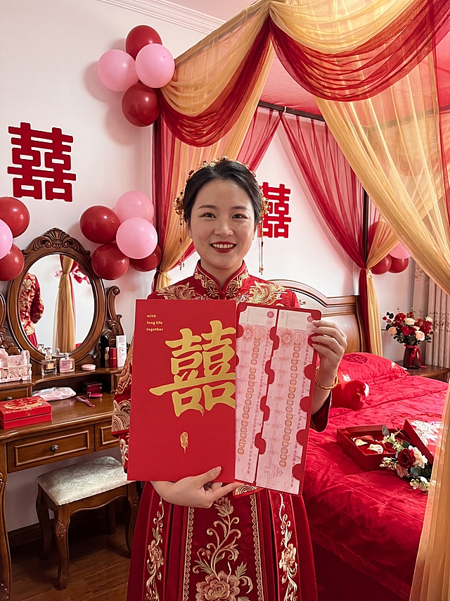 画面中心为身着红色中式传统礼服的人物，左手托住红包底部，右手扶在红包右侧边缘，将红包举在胸前，面部完整露出，笑容自然。 红包为左右两页展开式设计，占画面核心视觉区域近 50% 面积，保持红包原有样式不变。背景为布置温馨的婚房闺房，有红色喜字、气球、纱幔装饰，摆放着红色婚床、梳妆台、喜糖盒与花艺摆件，光线柔和温暖，充满家的氛围感。高清写实手机摄影风格，画面带有生活纪实感，质感清晰细腻，无过度磨皮或美颜，保留真实的皮肤纹理、手部细节、衣物刺绣质感，还原日常随手拍的真实氛围感，光影自然柔和。