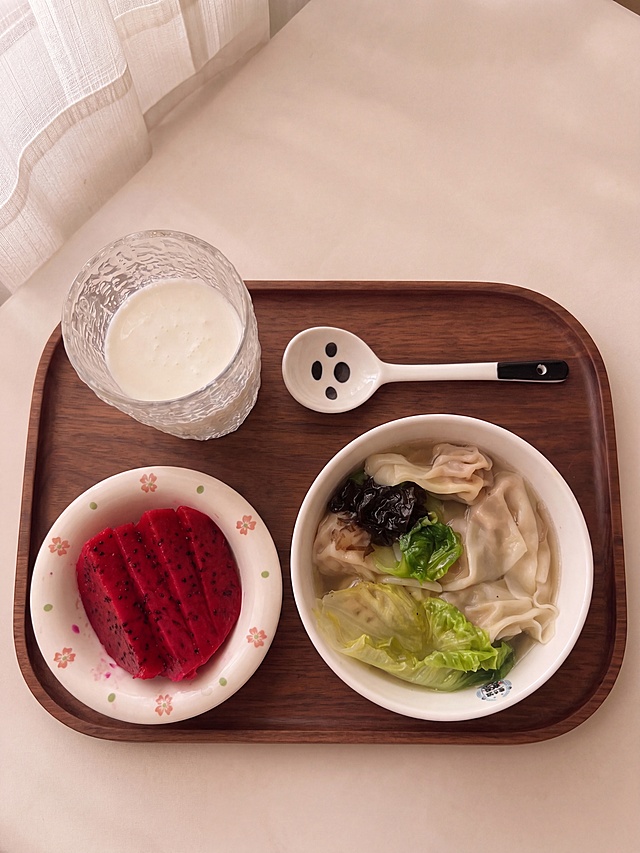 将图中的背景替换为image1中的米白色餐桌+深色圆角长方形胡桃木托盘+左上角白色纱帘的同一系列风格，保留图中的所有食物（馄饨汤、火龙果切片、牛奶杯、熊猫勺子）和餐具全部不变，整体光影和暖色调与image1保持完全一致，阴天窗边自然光效果，暖色系ins风格，氛围温馨治愈，俯视角度不变，看上去是同一个系列的早餐摆盘