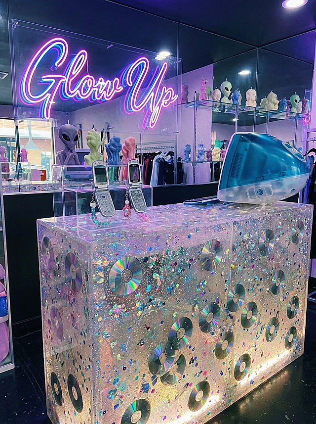 The checkout counter of a trendy, Y2K-themed lingerie store. The counter itself is a solid block of polished, translucent resin with glitter and old CD fragments embedded inside. Behind the counter, the wall features a large, cursive neon sign that says "Glow Up." Shelves behind the counter display quirky items like scented candles in funky shapes and retro flip phones. A vintage, bubble-shaped iMac G3 sits on the counter as a decorative piece. The area is cool, photogenic, and reinforces the brand's edgy personality.