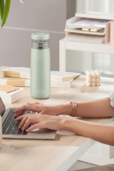 将办公室桌面场景中的粉色保温杯替换为浅绿色的ASD保温杯，保持保温杯的Logo、外形结构和尺寸比例完全不变，匹配场景中柔和的漫射光，在保温杯右侧生成自然柔和的投影，与场景光影完全一致，边缘过渡自然，整预览效果