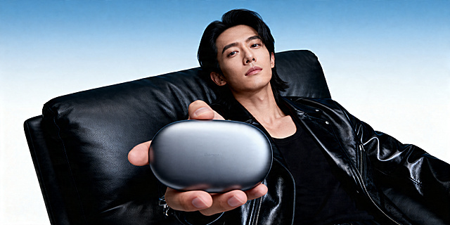 前景突出式中心构图，平视微仰视角，年轻亚洲男性模特，黑色中长发，黑色亮面真皮皮衣，黑色内搭，斜靠黑色超细腻牛皮革沙发，手掌向前托举椭圆形哑光金属智能设备，金属质感细腻高级；冷蓝白渐变背景，柔和精准布光，科技感光影，现代科技轻奢风格，数码产品商业宣传海报，构图聚焦产品，质感细腻，光影高级合理