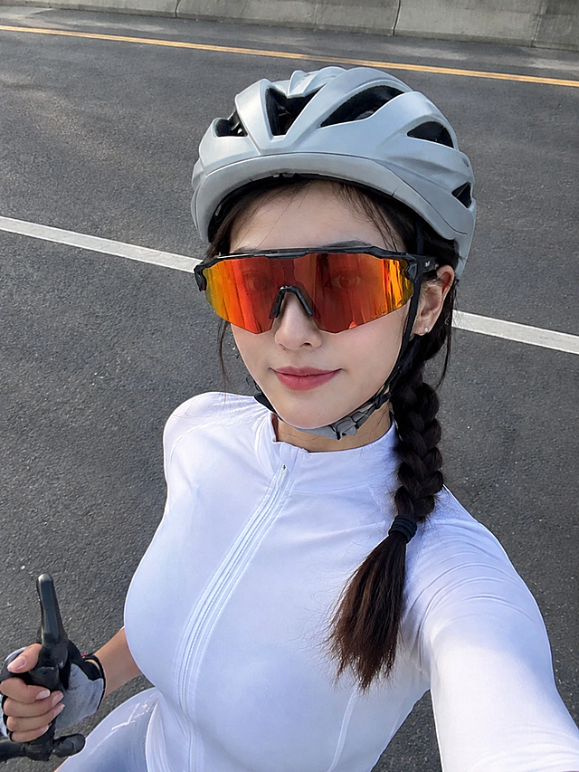 参考上一张生成的骑行女生自拍动作和构图，将女生佩戴的眼镜更换为用户新提供的这款骑行眼镜。严格保持新眼镜的所有几何特征、颜色、材质、镜框粗细、鼻托形状、镜腿样式完全不变：黑色镜框、红橙渐变反光镜片、一体式大镜片骑行风镜款式。漂亮的中国女生，五官精致，化着干净又漂亮的妆容，哑光底妆，穿着白色骑行服，佩戴浅灰色骑行头盔，背景为户外公路。无AI感，真实人像摄影，超高清，皮肤质感真实自然，光影自然。