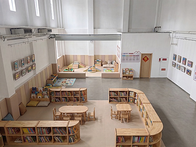 俯视室内二层实景摄影，保留原有建筑层高、顶部天窗、上层护栏、墙面管线、左右两侧展示挂画、带福字木门及门口宣传展板完全不变；结构改造：白色防护栏杆完整拆除，中间挑空镂空区域填平浇筑完整平整楼板，二层形成无落差连续完整平面；后方整面法治宣传墙体全部拆除打通，仅保留带木门区域原样不动；前景区域打造儿童阅览区：全圆角低矮原木绘本书架、无棱角儿童桌椅、加厚软包坐垫、多彩幼儿绘本，墙面做软包防撞护角，地面铺防滑环保地垫；后方打通开阔空间打造儿童活动区：全屋墙面全包防撞软包，搭配小型软包攀爬玩具、积木区、卡通软垫、绘本角，所有家具圆弧边角，无尖锐凸起，地面满铺防滑加厚EVA地垫；高处窗户自然光均匀柔和，保留原始白色墙面、工业风顶面管线，整体明亮温馨童趣，环保柔和配色，8K高清工装实拍质感，空间透视、建筑轮廓与原图完全匹配。