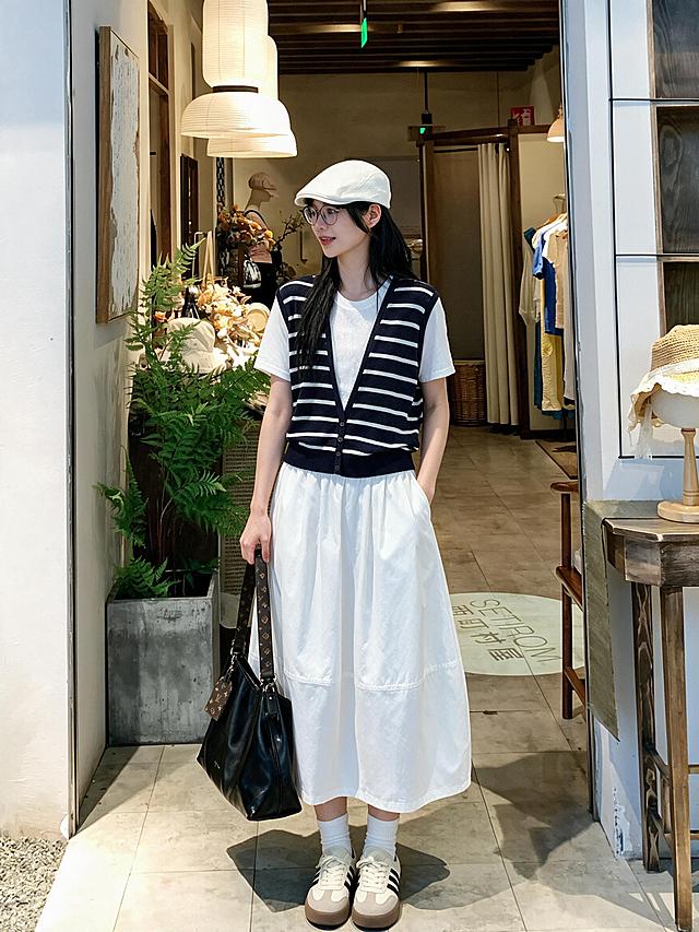 竖幅3:4，手机原生户外纪实人像，年轻东亚女生，黑色长直发，白色前进贝雷帽，细框眼镜，正面站立，一手插裙袋，一手拎包，日系简约文艺松弛气质。身穿黑白条纹针织假两件短袖上衣，外层V领针织，面料为细腻透气针织冰丝面料，清晰可见针织纱线肌理，织物带有细微网眼纹理，柔软垂顺。下装白色大摆茧型中长半裙，高腰松紧腰头，裙长至小腿中部，裙身蓬量饱满呈茧型弧度，裙身中下位置带有一条横向分割缝线，宽大圆润裙摆自然垂落，哑光梭织面料，带有柔和自然褶皱。脚上搭配白色中筒棉袜，脚穿米白拼浅灰德训板鞋，鞋身黑色三道条纹，厚棕底休闲鞋。手提黑色软质亮皮托特大包，搭配老花宽肩带与吊牌配饰。背景是日系文艺服装店门口，绿植盆栽，纸质灯笼，店内陈列服装，柔和漫射自然光，干净治愈日系小店氛围感，高清写实，完整保留衣物布料纹理细节，自然真实的光线，皮肤质感细腻通透，全身人像正面构图