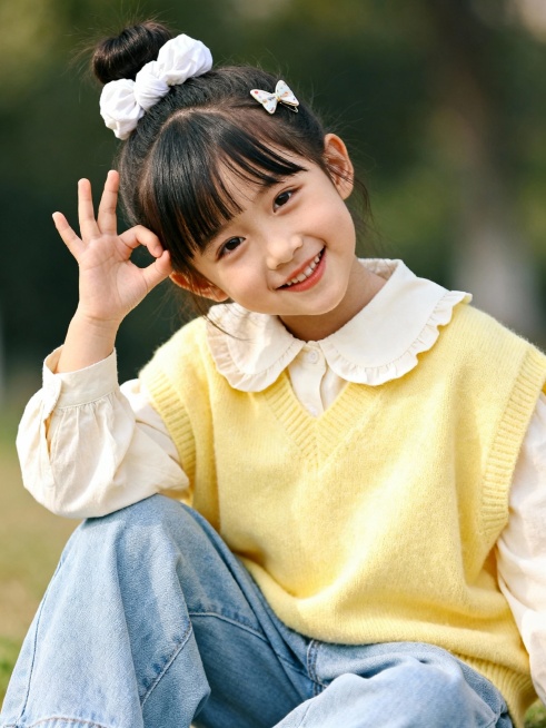 真实摄影影像，人物摄影，以为6岁的小女孩扎着丸子头，丸子头上带着白色布料的大发圈，前面斜刘海，刘海上夹着蝴蝶夹，穿着米白色娃娃领衬衫，柠檬黄宽松毛衣背心，浅蓝色宽松牛仔裤。微笑的看着镜头，头微微歪着，预览效果