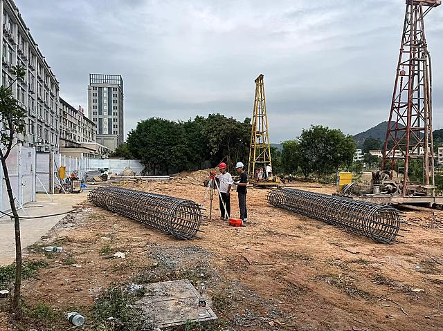 在这张建筑工地照片中，合理添加两个约5米长的圆柱形钢筋笼，参考第二张图的钢筋笼样式。钢筋笼放置在工地泥土地面上，位置自然合理，一个放在画面中景偏左区域，另一个放在画面右侧钻井设备附近的地面上。钢筋笼的透视、光影、阴影要与原图的阴天自然光环境完全一致，比例真实，与地面接触自然，融入场景不突兀，保持原图的写实摄影风格和画质，除添加钢筋笼外其余场景元素保持不变。