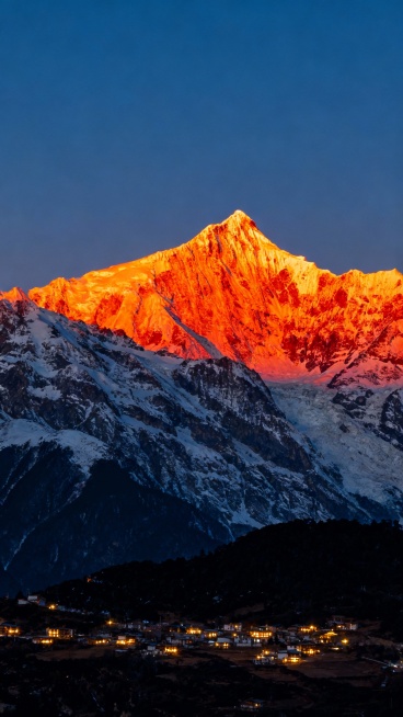 日照金山，壮丽雪山山顶被清晨阳光染成耀眼的金红色，山体覆盖皑皑白雪，山脚是灯火点点的宁静村庄，深蓝色渐变天空，写实风格，细腻光影，雪山纹理清晰，村庄灯光温暖，整体氛围神圣而静谧，不同角度展现雪山雄伟身姿与光影变化预览效果