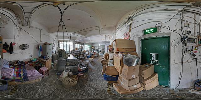 360度全景VR球形全景图，写实照片风格，食品加工小作坊内部场景，整体自然光室内光照，真实柔和光影，环境色调真实自然，照片质感，高清细节。画面中有清晰的安全出口标识和绿色安全出口门。画面中呈现多处火灾隐患点：1. 电线老化、乱拉乱接，电线外皮破损裸露，随意横跨天花板和墙面，走线混乱；2. 插线板超负荷串接使用，多个大功率电器同时插在串接的插线板上，插线板过载；3. 违规住人，角落放置床铺和生活用品，床铺紧邻加工设备和杂物堆放区；4. 货物和纸箱堵塞消防通道、堆放在安全出口门前，严重阻碍疏散通道。整体场景真实可信，细节丰富，具有警示意义。
