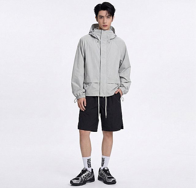 将图中模特的上衣替换为浅灰色连帽冲锋衣（图1款式），下装替换为带白色抽绳的黑色休闲短裤（图2款式），保持模特姿态和其他穿搭元素不变，服饰贴合身形自然