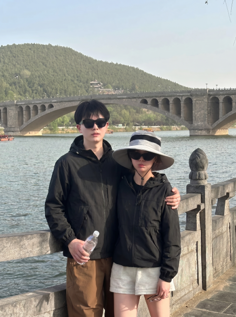 将图2的黑色连帽外套分别穿到图1河边护栏这对情侣身上，男女都换上这件黑色外套，替换掉原来的黑色T恤，过渡自然平整，保持原来的大桥河景背景和姿势，图片比例3比4预览效果