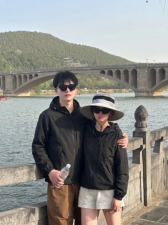 将图2的黑色连帽外套分别穿到图1河边护栏这对情侣身上，男女都换上这件黑色外套，替换掉原来的黑色T恤，过渡自然平整，保持原来的大桥河景背景和姿势，图片比例3比4