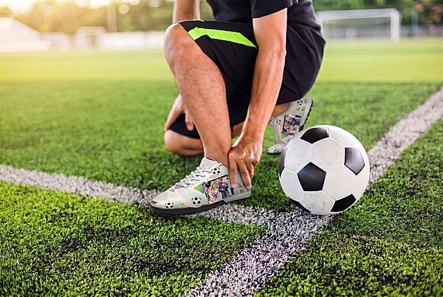 参考图1的风格和构图，将图2这款米白底定制足球鞋放在足球场草地上，人物单膝跪地手扶脚踝，旁边放一个足球，保留图2足球鞋原有的米白底、足球几何图案和婚礼合影印花，真实户外摄影，暖调阳光质感，和图1风格一致