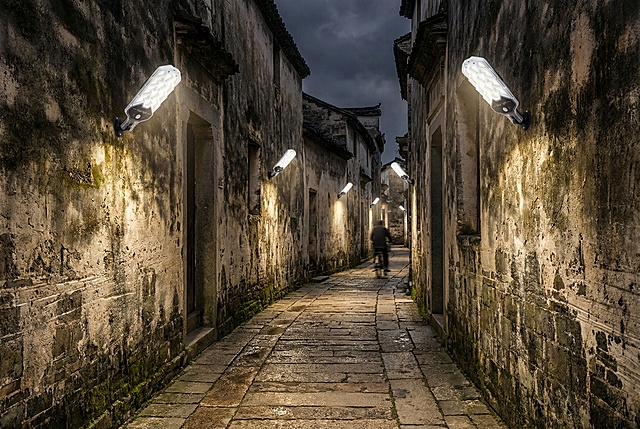 一条蜿蜒的窄巷，两侧是墙壁。在墙面约5米高的位置，等距安装着5-6盏图上的太阳能壁灯。灯光是白光向下洒在古老的石板路上，完美勾勒出小巷的深邃感和历史韵味。远景处可模糊看到一辆自行车经过。