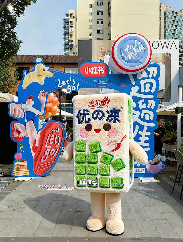 以优の冻仙草冻饮料包装盒为整体外形，做成Q版可爱的人偶玩偶服，参考小红书毛绒玩偶的短毛绒质感，材质柔软蓬松，面部圆润可爱，有圆圆的大眼睛和粉色腮红，整体造型生动，细节清晰，户外街道场景