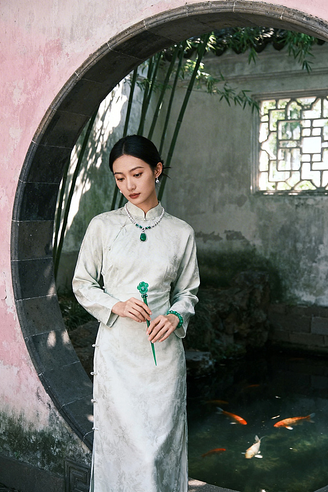 视觉关键词：东方美学、静谧深远、人文意境
场景描述：
模特身穿素雅中式长裙或改良旗袍，立于苏州园林的月洞门旁，或倚靠徽派建筑的木雕窗棂。手中轻持一枚翡翠玉簪或白玉手镯，目光低垂，姿态从容。背景可见斑驳粉墙、疏竹影斜、浅水池鱼，自然光线从窗格透入，勾勒珠宝温润光泽。整体色调以青灰、墨绿、暖白为主，突出“人在景中，玉在光中”的沉浸感。

镜头建议：中景至全景，注重环境与人物的构图平衡，焦点在珠宝与模特眼神的交汇处。