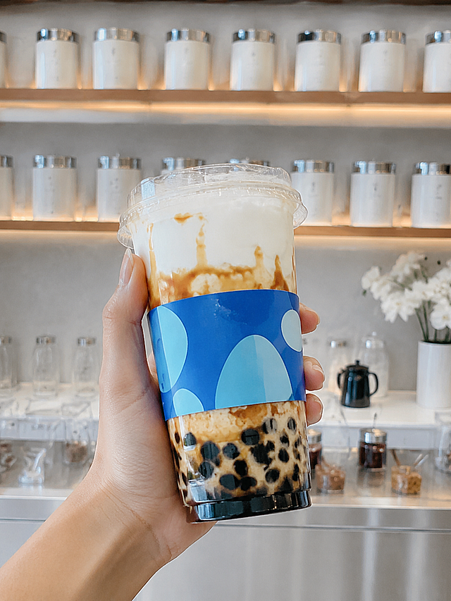 竖版小红书奶茶种草截图，画面主体手部手持透明塑料奶茶杯，杯身蓝色多巴胺杯套，无logo，杯内黑糖珍珠、奶盖与焦糖挂壁分层；背景奶茶店内货架整齐摆放，干净背景，高清写实，8K
