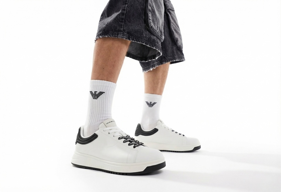 以参考图的构图、人物穿搭、拍摄角度为基础，将图中的运动鞋替换为image2的白黑配色休闲板鞋，在人物穿着的白色袜子上添加image3的Emporio Armani黑色logo，保持画面为纯白背景，风格预览效果