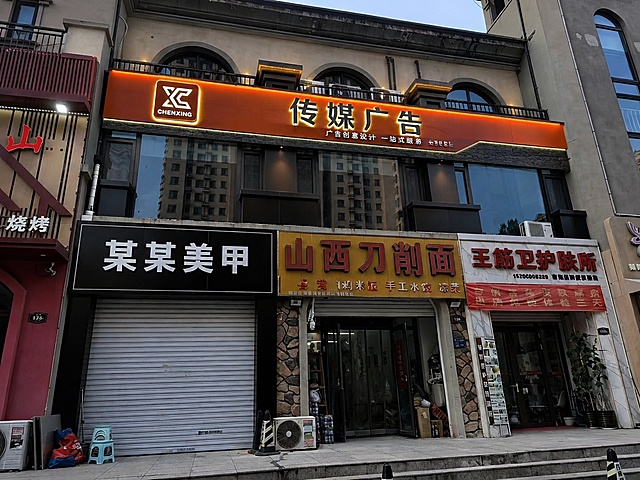 将图二中上层黑色的"尘星传媒"门头区域替换为高端广告公司门头设计，高大上简约爱马仕橙系列风格。门头左侧是亚克力转角灯箱logo，主文字"传媒广告"为背发光文字效果，下方小字"广告创意设计 一站式服务"为无边字工艺。门头底板采用两种以上材质拼接设计，上层为爱马仕橙色哑光金属板，下层为深灰色石材纹理面板，材质对比鲜明。整体风格现代简约、高端大气，爱马仕橙为主色调搭配深灰色和金属线条点缀，夜晚灯光效果柔和高级，商业门头实景拍摄质感，超清细节，专业建筑摄影风格。保持图二原有建筑结构、下层店铺和周边环境不变，仅替换上层黑色门头部分，透视和光影与原图一致。