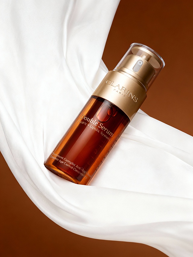 奢华高级的商业美妆摄影风格，高质感精致渲染；画面采用暖棕与柔白交织的高级暖调，整体氛围优雅奢华且富有细腻质感；主体倾斜摆放于画面中间白色流动的缎面的褶皱之上；白色缎面的柔滑质感既承托产品，细腻褶皱强化奢华优雅氛围。上下透出背后棕色背景