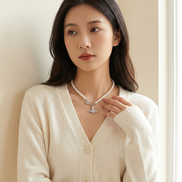 [主图 2] 年轻亚洲女性佩戴西太后满钻土星珍珠锁骨项链中景展示。[氛围风格] 高级温柔氛围感，生活化时尚实拍风。[画质] 商业摄影质感、光线柔和通透、产品清晰真实、画面干净高级。[元素] 同一位年轻女性模特，穿着米白色针织开衫，站在极简浅奶白色墙面背景前，自然侧身站立，手轻轻搭在锁骨位置，完整展示项链的佩戴整体效果。保持项链外观（即物理结构、材质质感、颜色）与输入产品图绝对一致，杜绝任何变形和色差，精细化处理。商品主体展示清晰，无任何遮挡。画面满屏无边框。[光影] 柔和落地窗自然光，均匀明亮，珍珠和钻石的光泽自然通透，整体画面干净明亮。[配色] 主色调浅奶白，搭配服装米白，突出项链银白钻闪。[视角构图] 胸部以上平视中景构图，人物居中，景深适中。[文字版式] 无添加文案，纯画面展示。[比例分辨率] 1:1 比例，2k 分辨率；画面超高清。[负向提示词] 变形，水印，文字错乱，产品图模糊，背景杂乱，换模特换脸，服装不一致。